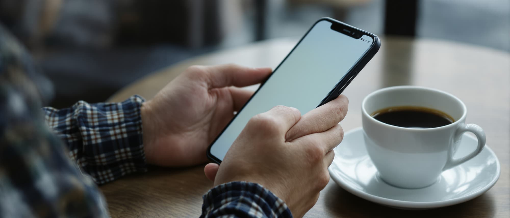 Hands holding a phone at a café table beside a cup of black coffee, the screen still blank.